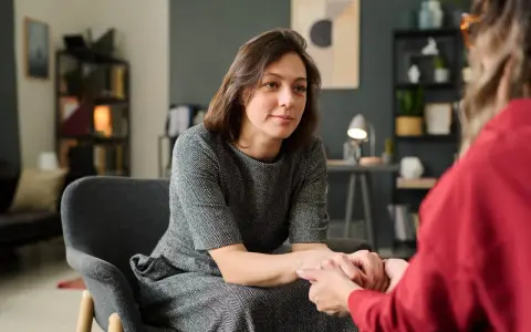 Kobieta w szarej sukience siedzi w fotelu, trzymając dłonie drugiej osoby. To scena wspierającej terapii gestalt.