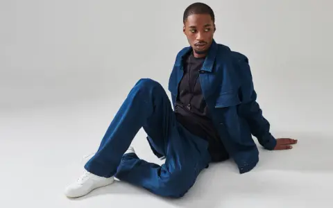 Giovane uomo con completo in materiale jeans, seduto a terra, con giacca e pantaloni oversize.