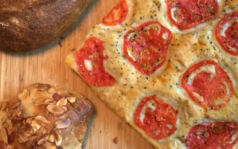 Focaccia receta con tomates frescos y hierbas, junto a un pan rústico y un bollo con almendras.