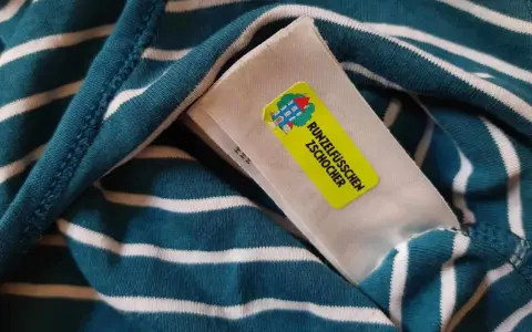 Namensetiketten Kita Erfahrungen: Ein gestreiftes Shirt mit einem gelben Aufkleber "Runzelfüßchen Zschocher" und einem Hausmotiv.