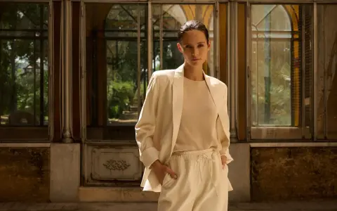 Mujer con blazer beige y pantalones de vestir, de pie frente a una estructura con ventanas.