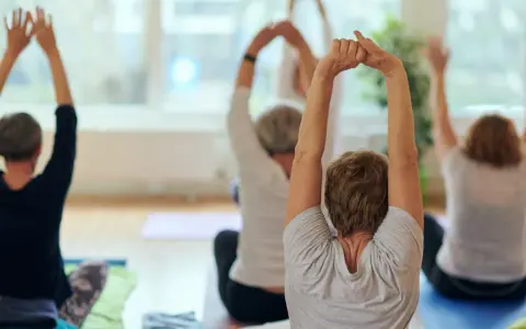 Grupa seniorów ćwiczy jogę, podnosząc ręce do góry. Wszyscy siedzą na matach, skupieni na ruchu.
