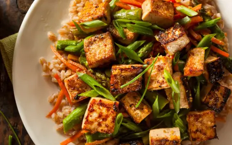 Talerz pełen brązowego ryżu z grillowanym tofu, szparagami i marchewką, posypany szczypiorkiem.