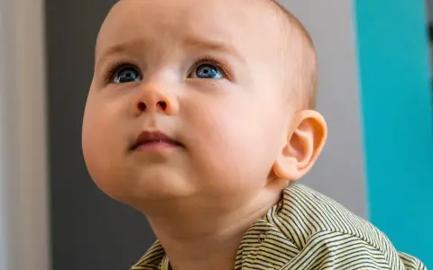 Occhi azzurri di un neonato di 10 mesi che guarda in alto, con un'espressione curiosa. Indossa una maglietta a righe gialle e nere.