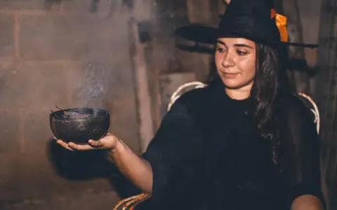 Mujer con disfraz bruja casero adulto, con sombrero negro y naranja, sosteniendo un cuenco humeante.