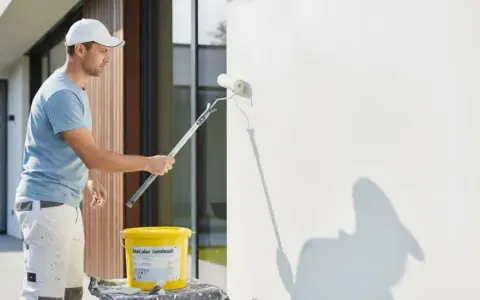 Mann streicht Fassade mit weißer Farbe. Für ein gleichmäßiges Ergebnis ist eine Grundierung notwendig.