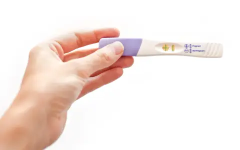 Dłoń trzyma test ciążowy z wynikiem pozytywnym. W okienku widoczny jest symbol plusa, oznaczający ciążę.