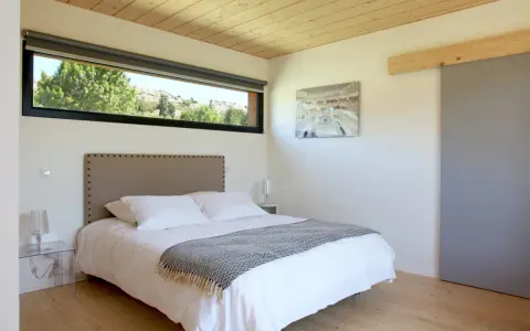 Chambre couleur taupe avec un grand lit blanc, une tête de lit capitonnée et une vue sur la nature par la fenêtre.