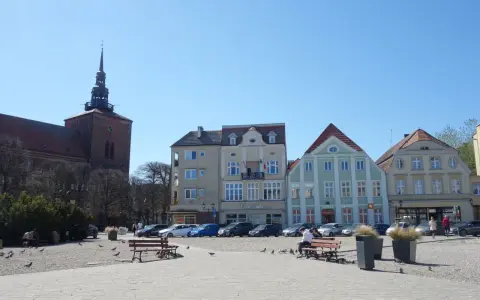 Słupsk, stary rynek: kościół z wieżą, kolorowe kamienice, ławki, ludzie i gołębie na brukowanym placu.