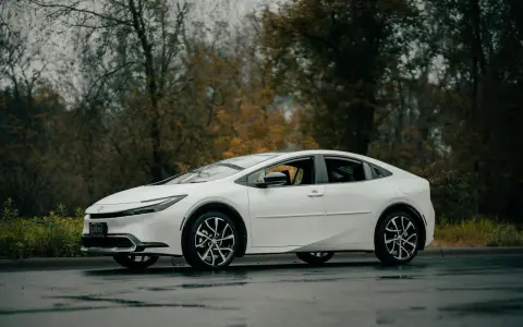 Biała Toyota Prius zaparkowana na mokrej drodze, z drzewami w tle.