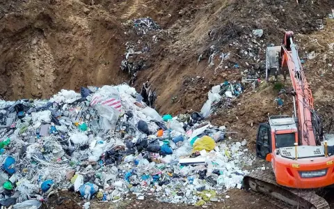 Ogromna góra śmieci, tworząca nielegalne wysypisko, z koparką w tle. Widać worki, folie i różne odpady.