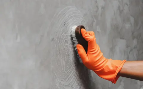 Mano con guante naranja limpiando pared gris con cepillo, aprendiendo como limpiar las paredes de color sin dañar la pintura.