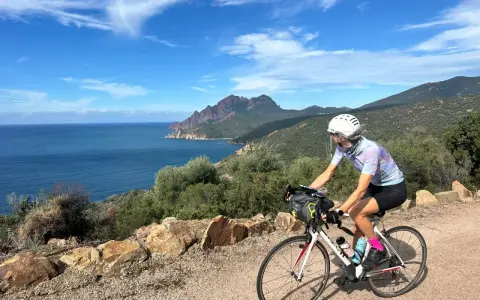 Ciclista pedala lungo la costa rocciosa della Corsica, con il mare blu e montagne sullo sfondo. Un'avventura in bici indimenticabile.