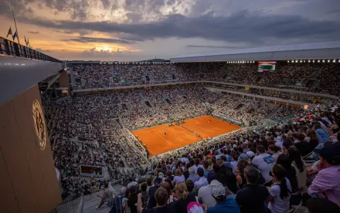 Widok na pełny stadion podczas meczu na kortach ziemnych. Zachodzące słońce oświetla trybuny pełne widzów, tworząc atmosferę emocjonującego **Roland Garros live**.