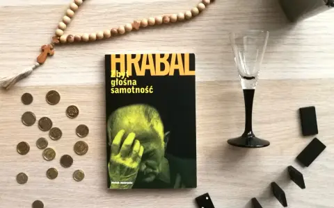 Koraliki różańca, monety, kieliszek i domino otaczają książkę "Zbyt głośna samotność".