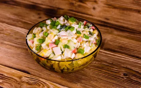 Pyszna sałatka z surimi, z kukurydzą, jajkiem i szczypiorkiem, podana w szklanej misce na drewnianym blacie.