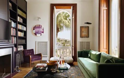 Soggiorno elegante in una casa stile Liberty, con divano verde, poltrona viola, libreria e vista panoramica.