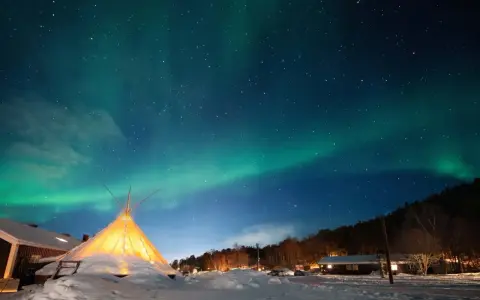 Nocne niebo nad ośnieżoną wioską, gdzie nie zachodzi słońce. Tańcząca zorza polarna nad oświetlonym tipi i domkami.