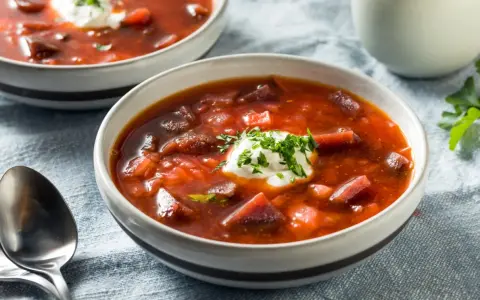 Dwie miseczki aromatycznej zupy z botwiny na mięsie, ozdobione kleksem śmietany i posiekaną natką pietruszki.