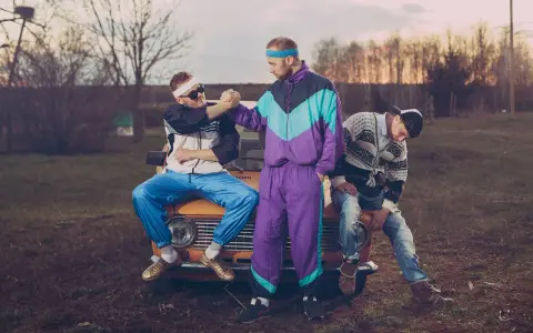 Trzech mężczyzn w dresach i okularach przeciwsłonecznych, uosabiających styl lat 90., siedzi na masce starego samochodu.