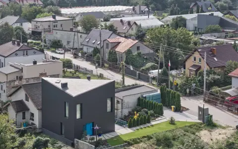 Nowoczesny, czarny dom o trójkątnym kształcie wyróżnia się na tle tradycyjnych terenów mieszkaniowych b. Otoczony zielenią, z niebieskimi drzwiami.