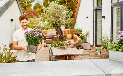 Mann hält Topf mit lila Blumen, bereit, die Dachterrasse zu bepflanzen. Eine Frau sitzt am Tisch und arbeitet im Garten.