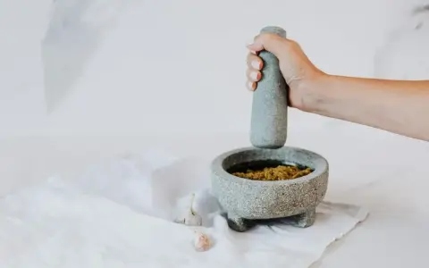 Dłoń uciera w moździerzu aromatyczne pesto z bazylii. Obok leżą ząbki czosnku.
