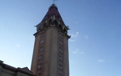 Wysoka wieża z czerwonym dachem, przypominająca dom rybaka, na tle błękitnego nieba.