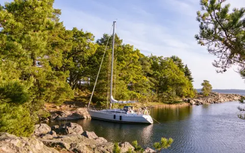 Segelboot liegt ruhig im Wasser, bereit, den Anker zu legen oder zu setzen. Grüne Bäume und Felsen säumen die Küste.