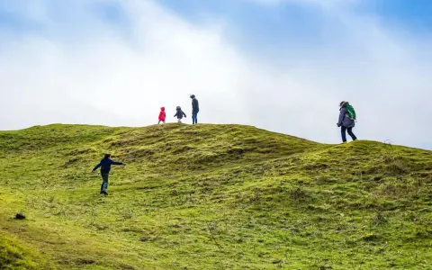 Famiglia esplora sentieri per bambini su una collina verdeggiante sotto un cielo azzurro e nuvoloso.