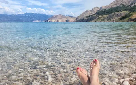 Stopy w klapkach na kamienistej plaży w **Krk National Park**. Woda krystalicznie czysta, w tle góry.