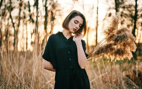 Młoda kobieta w czarnej sukience pozuje na tle zachodzącego słońca i trzcin. Idealna modelka do nauki kurs fotografii portretowej.