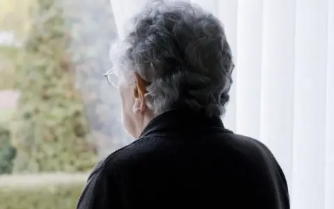 Starsza pani z siwymi włosami patrzy przez okno. Czeka na emeryturę po mężu, wspominając wspólne chwile.