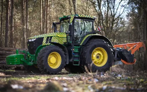 Zielony traktor John Deere z pomarańczowym mulczerem, gotowy do pracy w lesie.
