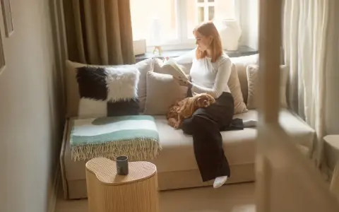 En kvinna läser en bok i en liten bäddfåtölj med sin hund i knät. En grön och vit pläd ligger över fåtöljen.
