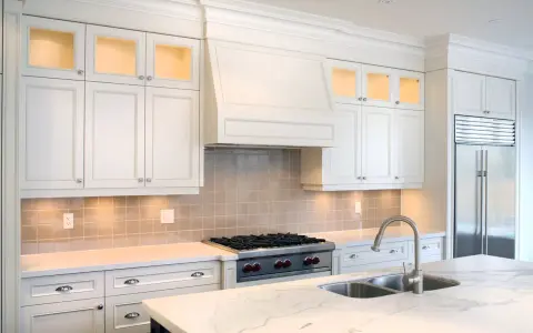 Cocina moderna con campana extractora decorativa sobre estufa. Gabinetes blancos, encimera de mármol y refrigerador de acero inoxidable.