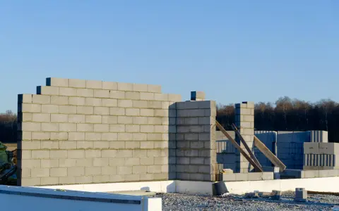 Budowa domu w toku. Wzniesiona ściana nośna z bloczków betonowych, w tle kolejne elementy konstrukcji i las.