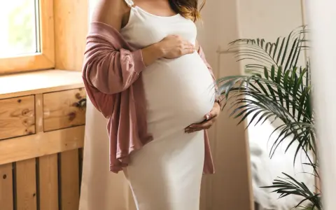 Frau in weißem Kleid und rosafarbenem Cardigan hält ihren Bauch, 34 ssw. Ein Fenster und eine Pflanze sind im Hintergrund.