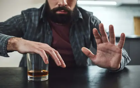Mężczyzna z zarostem odpycha szklankę z alkoholem, walcząc z chorobą alkoholową. Gest dłoni symbolizuje odmowę i chęć zmiany.