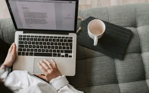 Person arbetar på laptop i soffan, en kopp står bredvid. Perfekt för dig som söker bra utbildningar på distans.