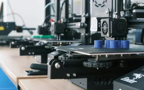 Drukarki 3D pracują, tworząc niebieskie cylindry. W tle widać kolejne maszyny, a na biurku leży smartfon, gotowy do zarządzania drukiem przez Raspberry Pi z OctoPrint.
