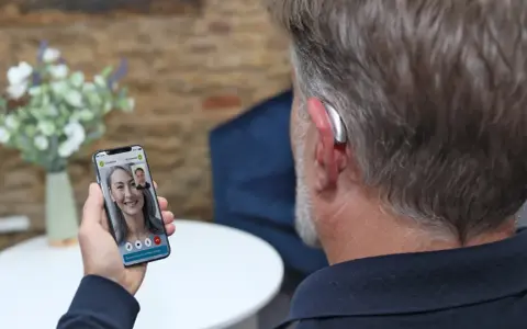 Mężczyzna z aparatem słuchowym rozmawia przez wideorozmowę na telefonie. Aplikacja na telefon pozwala na zdalne dopasowanie aparatów.