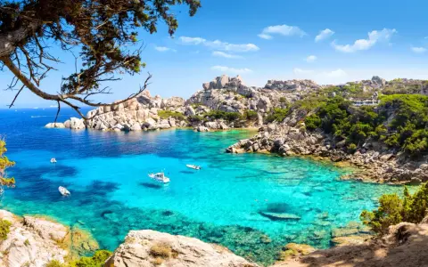 Turkusowa woda i skaliste wybrzeże Costa Smeralda na Sardynii. Kilka łodzi kołysze się na falach.