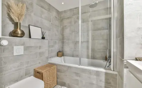 Salle de bain entierement carrelée de gris, avec baignoire, douche, lavabo et WC. Un panier en osier et des herbes séchées apportent une touche naturelle.