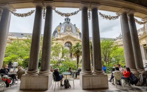 Vue sur le Grand Palais depuis un portique, avec des visiteurs profitant d'un café dans le jardin. Un petit palais, un grand palais.