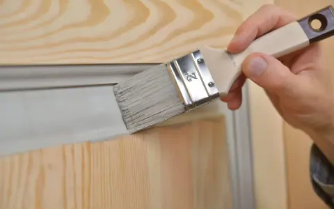 Une main utilise un pinceau pour relooker une porte avec des moulures, appliquant une peinture grise sur le bois clair.