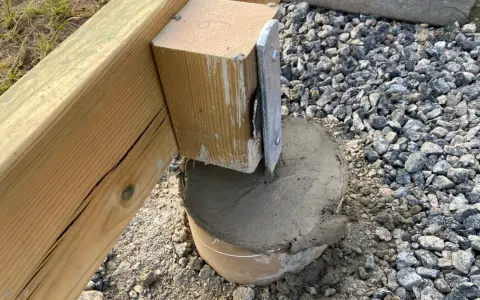 En trästolpe fästs i en betongplint, en del av arbetet med att bygga altan på plintar. Grus och jord syns runt plinten.