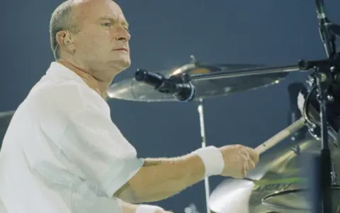 Phil Collins, w białej koszuli, gra na perkusji. Legendarny muzyk, znany z zespołu Genesis.