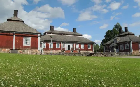 Malingsbo herrgård med sina röda trähus och stenfundament, omgivet av en grönskande äng med vita blommor under en blå himmel med moln.