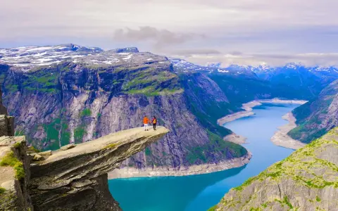 Två personer står på Trolltunga, en av Norges mest ikoniska och fina platser. En dramatisk klippavsats med en turkos sjö nedanför och snöklädda berg i bakgrunden.